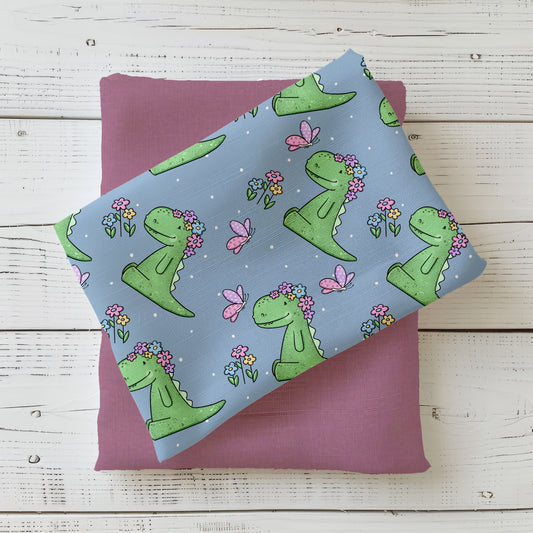 Mockup gefalteter Stoff mit dem Design „Rexeline“ von Katzengold: Liebevoll gestalteter Dino-Stoff mit Blumen und Schmetterlingen auf hellblauem Hintergrund – perfekt für Baby- und Kindermodelle.