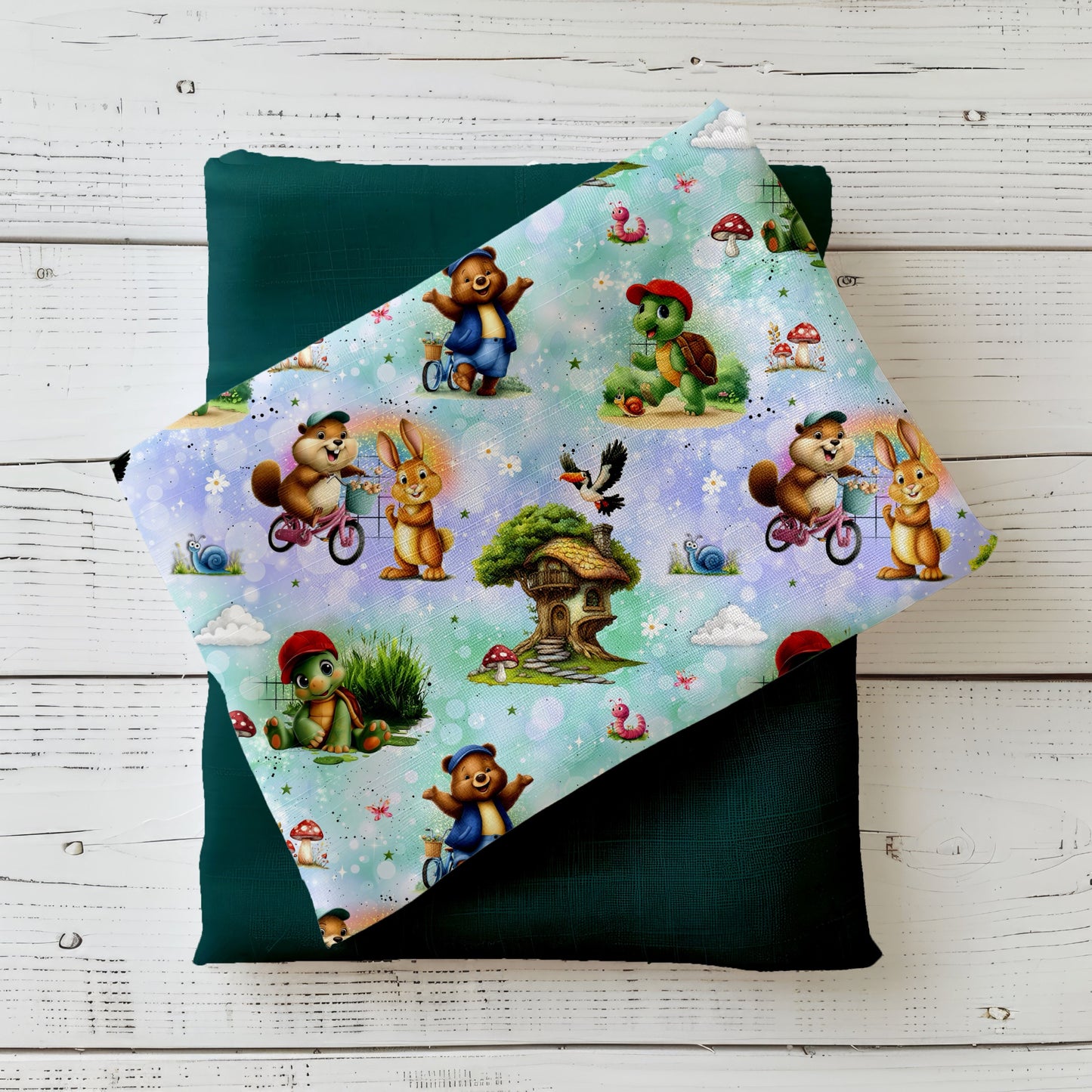 Mockup: Stoff „Schildi & Freunde“ mit kleiner Schildkröte als Hauptfigur, Bär, Hase und Biber (teils auf dem Fahrrad), Baumhaus, Pilzen, Schnecken und Wolken auf funkelndem Bokeh-Hintergrund; aufgelegt auf dunkelgrünem Kombistoff, fotografiert auf hellem Holz.