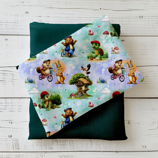 Mockup: Stoff „Schildi & Freunde“ mit kleiner Schildkröte als Hauptfigur, Bär, Hase und Biber (teils auf dem Fahrrad), Baumhaus, Pilzen, Schnecken und Wolken auf funkelndem Bokeh-Hintergrund; aufgelegt auf dunkelgrünem Kombistoff, fotografiert auf hellem Holz.