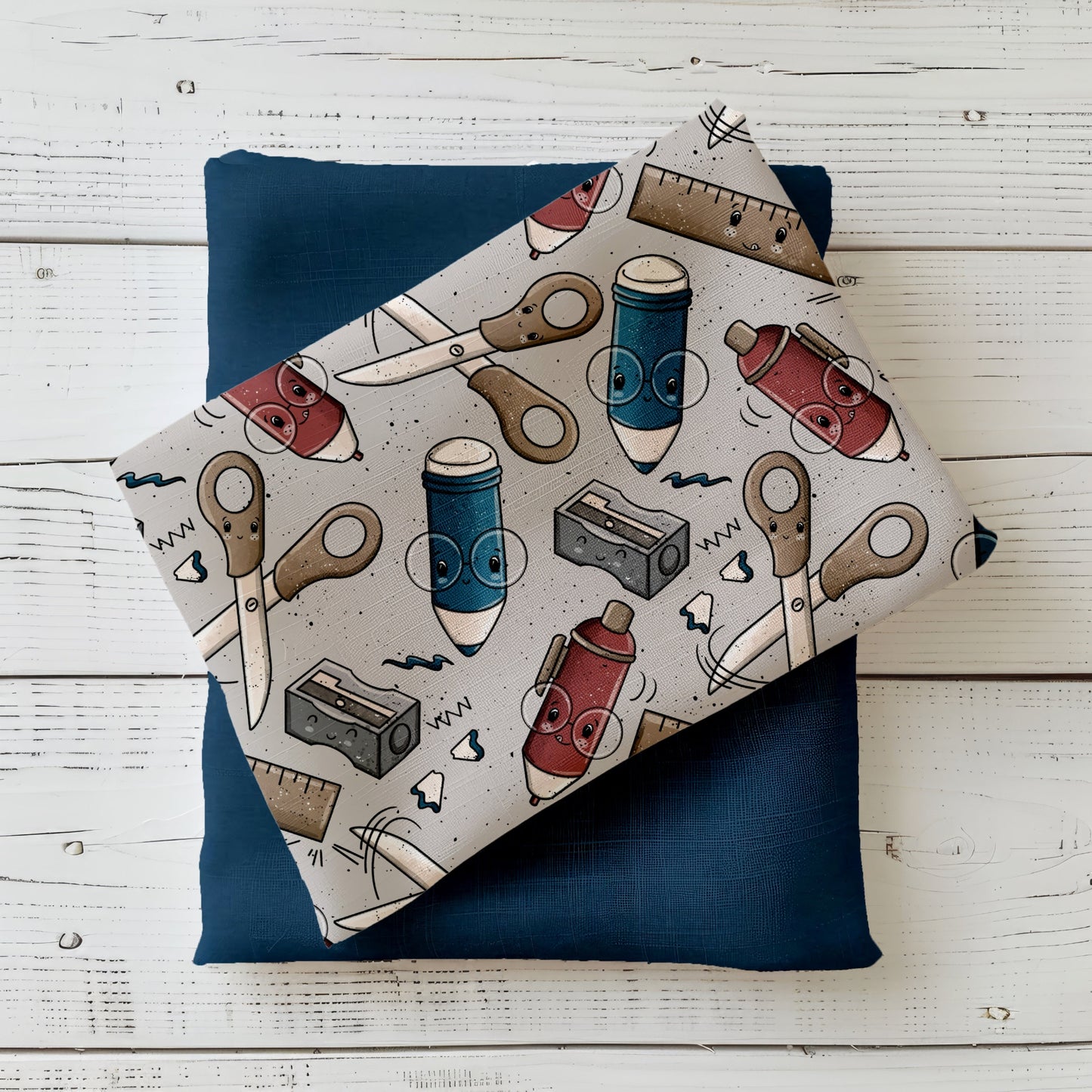 Stoff-Mockup mit dem Design „Schreibzeug – Grau“: grauer Stoff mit gleichmäßig verteilten Motiven wie Scheren, Stiften, Linealen und Spitzern in Beige-, Blau- und Rottönen. Der Stoff ist locker gefaltet arrangiert und mit einem dunklen Uni-Stoff kombiniert, um Farbwirkung, Motivgröße und Kombinationsmöglichkeiten der Meterware realistisch darzustellen. Design von Katzengold-Designs.