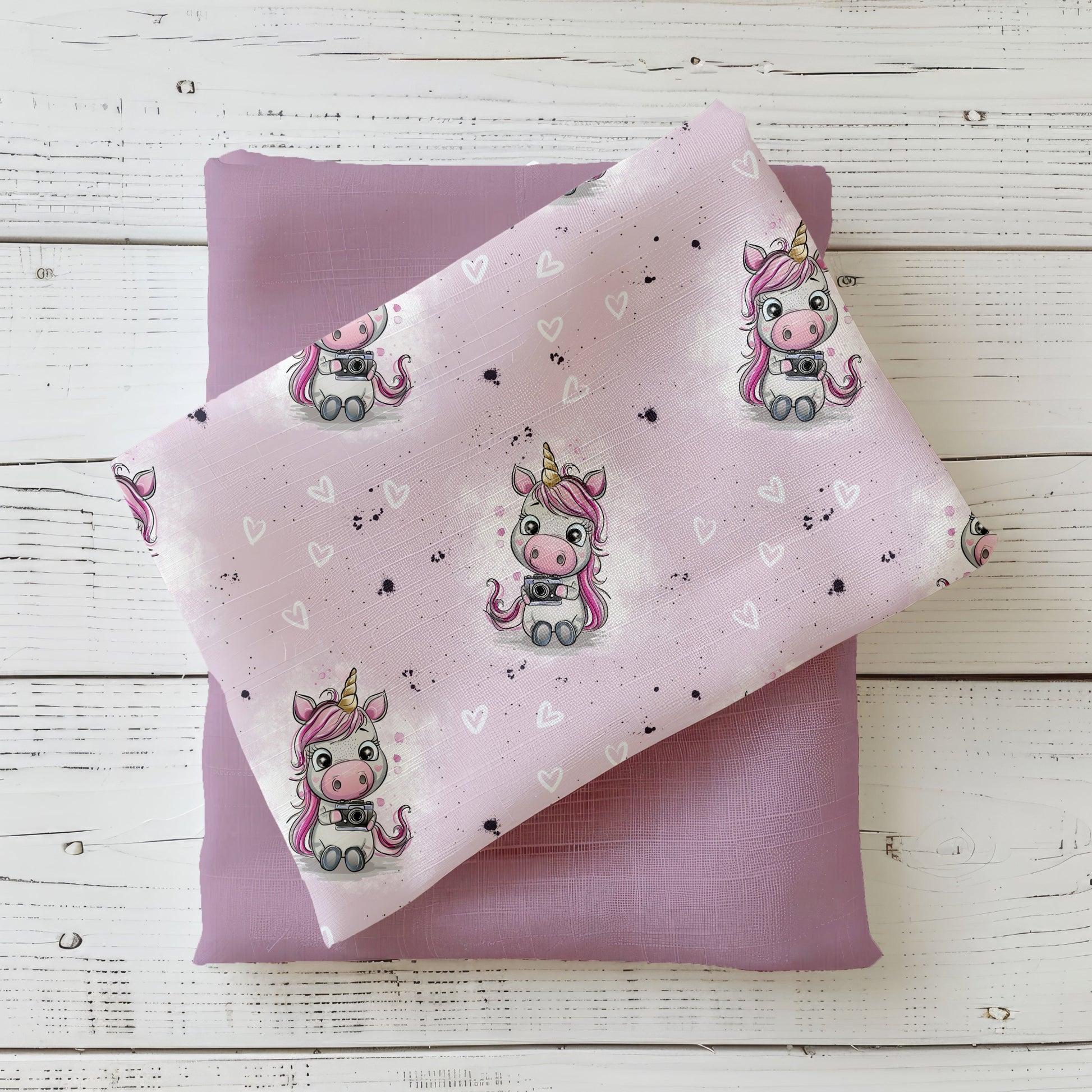 Rosa Einhornstoff „Smile“ mit Herzchen und Kamera-Einhorn, dekoriert als Stoffstapel auf violettem Untergrund.