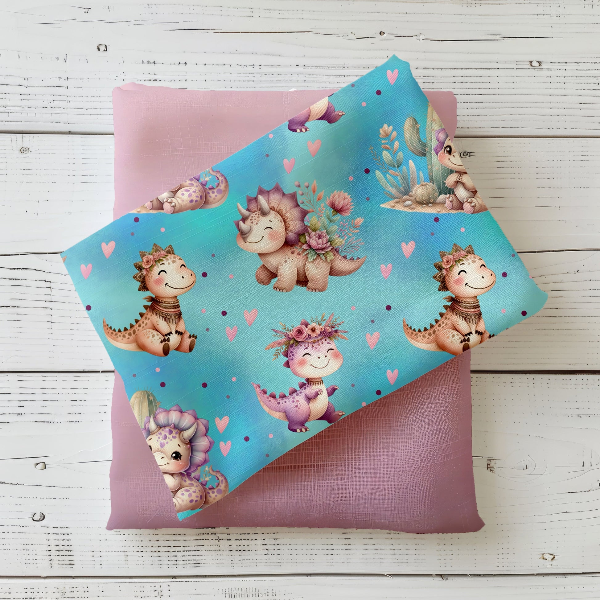 Mockup mit gefaltetem Stoff „Süße Dinos“ auf mintblauem Hintergrund mit niedlichen Dinosauriern, Herzen und Blumen, arrangiert auf einem rosa Uni-Stoff auf weißer Holzfläche.
