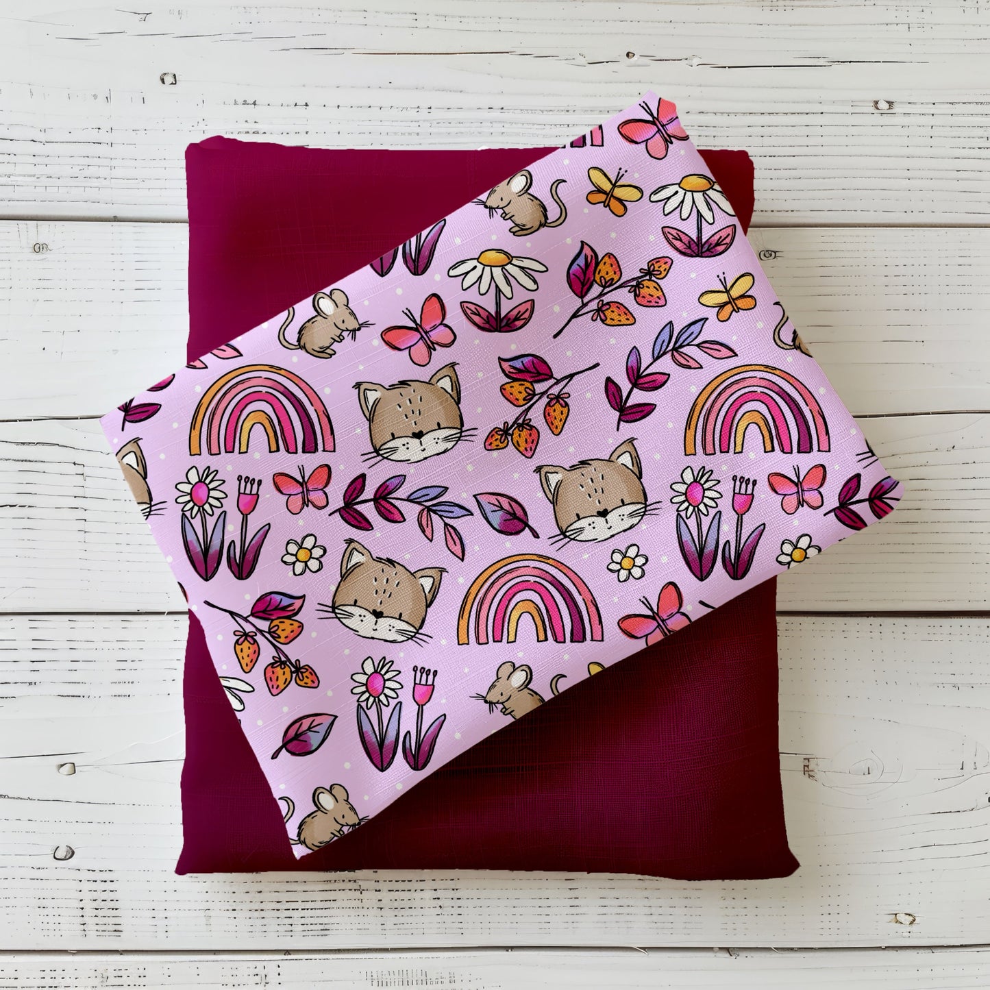 Bunter Stoff mit Regenbögen, Blumen und niedlichen Tiermotiven, auf einem Kissen drapiert, in kräftigen Rosa- und Beeretönen mit sommerlicher Ausstrahlung.