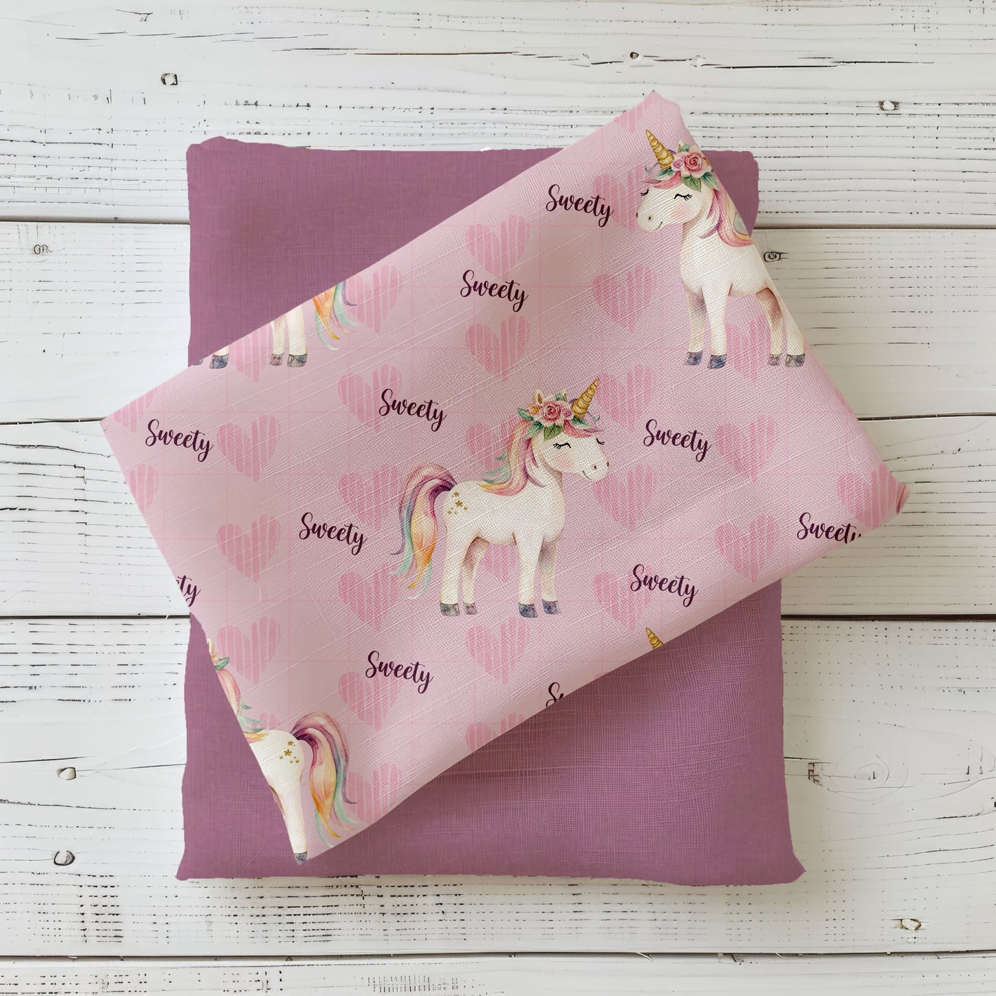 Mockup mit rosa Einhornstoff Sweety: gefalteter Stoff mit weißem Einhorn, Regenbogenmähne, Blumenkranz und rosa Herzchen auf Holzuntergrund – kombiniert mit passendem Uni-Stoff in Flieder.