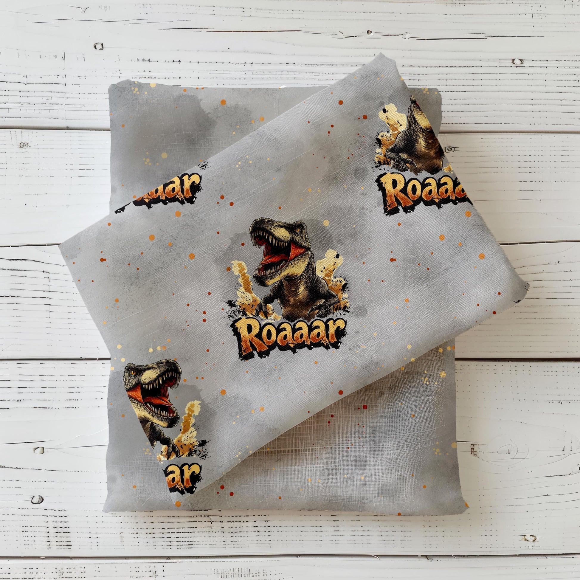 Mockup eines grauen Dino-Stoffs mit großem brüllenden T-Rex und Roaaar-Schriftzug, dekorativ auf einem dunkelgrünen Kombistoff drapiert – fotografiert auf hellem Holzuntergrund.