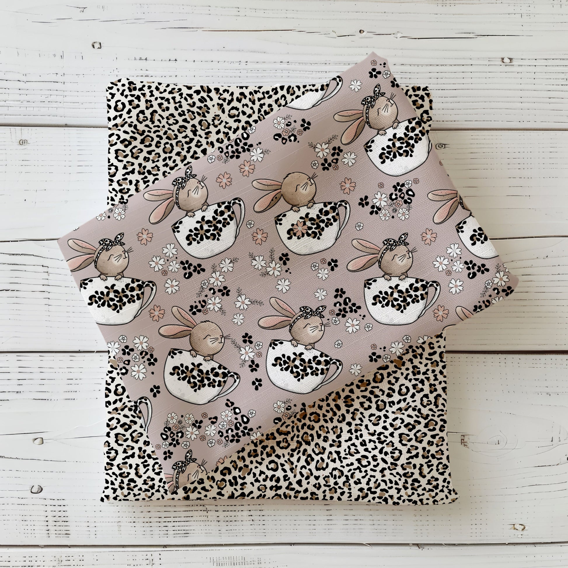 Stoffdesign „Tassenhase“ mit niedlichen Schlappohren-Häschen in Tassen, Blüten und Leo-Mustern in sanften Beige- und Rosétönen, fotografiert als gefaltetes Stoff-Mockup auf hellem Holzhintergrund.