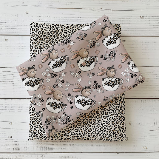 Stoffdesign „Tassenhase“ mit niedlichen Schlappohren-Häschen in Tassen, Blüten und Leo-Mustern in sanften Beige- und Rosétönen, fotografiert als gefaltetes Stoff-Mockup auf hellem Holzhintergrund.