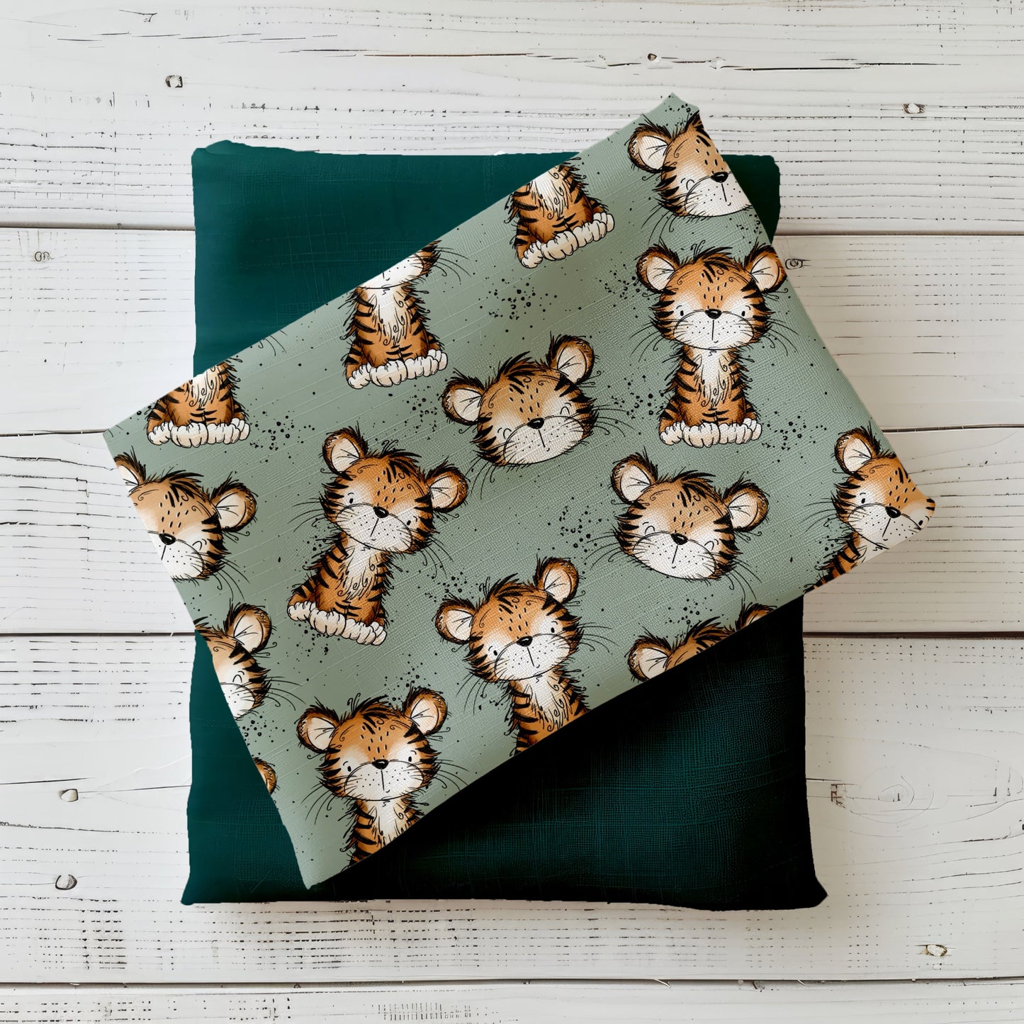 Stoff-Mockup mit liebevoll illustriertem Tigerdesign auf salbeigrünem Hintergrund. Der Stoff ist locker gefaltet arrangiert und mit einem passenden Uni-Kombistoff in dunklem Grün kombiniert. Die niedlichen Tigerköpfe und sitzenden Tiger sind gleichmäßig über die Fläche verteilt, ergänzt durch feine Sprenkel. Das Mockup zeigt die Wirkung des Designs im Nähkontext und vermittelt Farbwirkung, Motivgröße und Kombinierbarkeit der Stoffmeterware. Design von Katzengold-Designs.