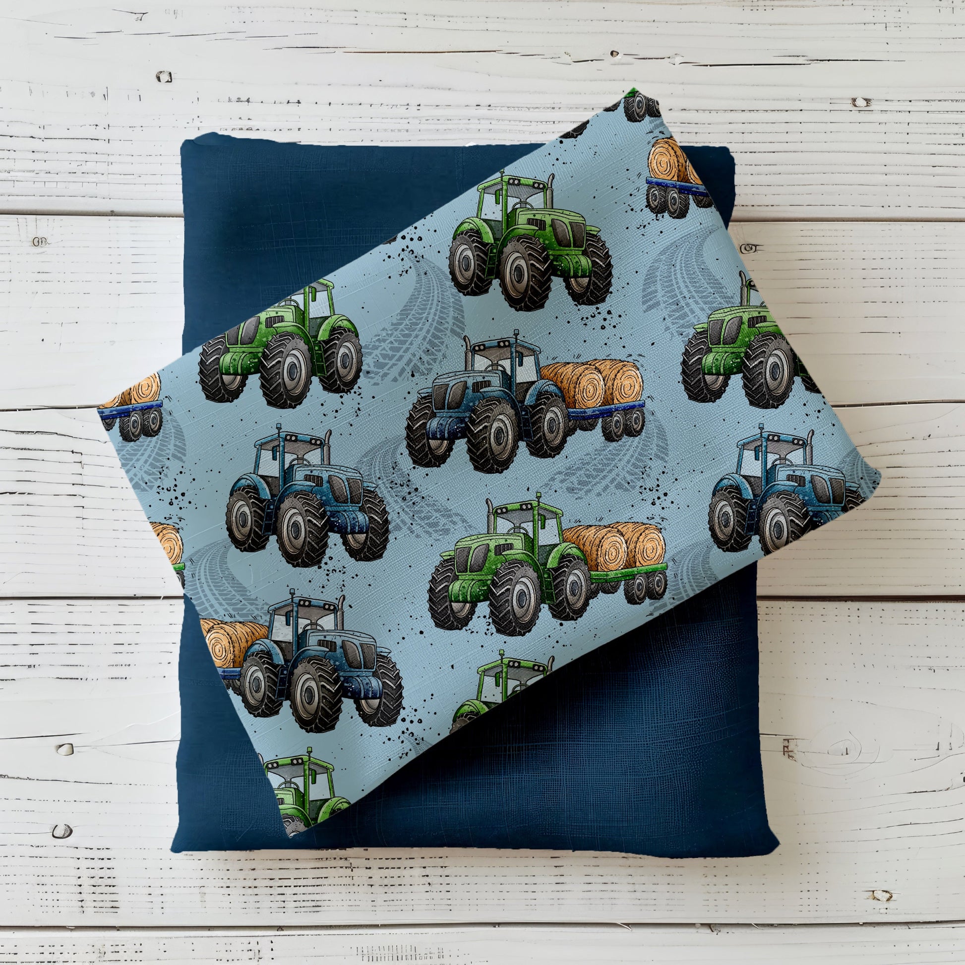 Mockup: Stoff „Trecker-Crew“ mit grünen und blauen Traktoren sowie Strohballen-Anhänger auf hellblauem Hintergrund mit Reifenspuren; aufgelegt auf dunkelblauem Kombistoff, fotografiert auf hellem Holz.