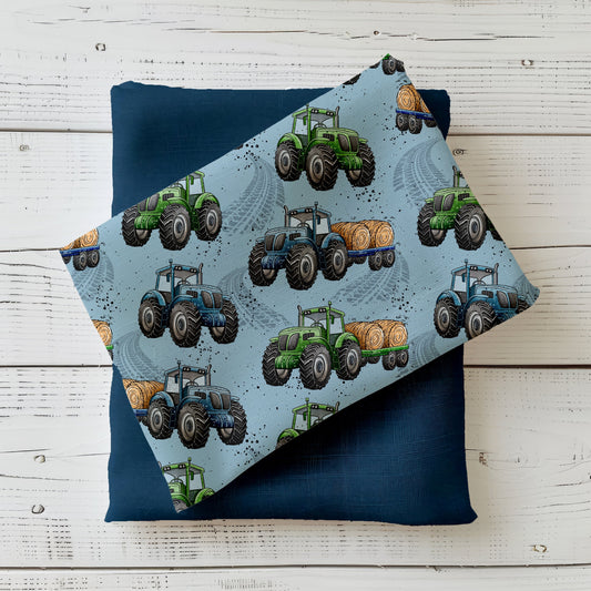 Mockup: Stoff „Trecker-Crew“ mit grünen und blauen Traktoren sowie Strohballen-Anhänger auf hellblauem Hintergrund mit Reifenspuren; aufgelegt auf dunkelblauem Kombistoff, fotografiert auf hellem Holz.