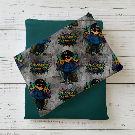 Mockup eines gefalteten Stoffes mit dem „Trouble Maker“-Design: Wiederholendes Graffiti-Muster mit coolem Jungen und buntem Schriftzug auf grauer Betonwand – ideal für modische Kinderkleidung im Streetwear-Stil.