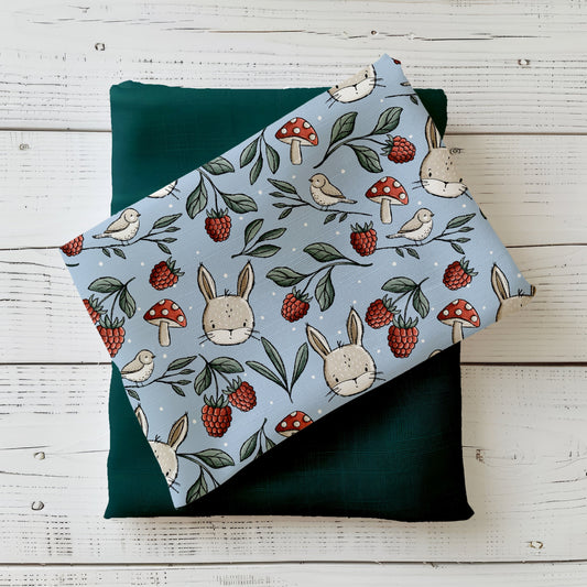 Gefalteter „Wiesentag“ Stoff von Katzengold-Designs – Häschen, Beeren, Pilze und kleine Vögel auf blaugrünem Hintergrund, dekorativ auf dunklem Grünton präsentiert
