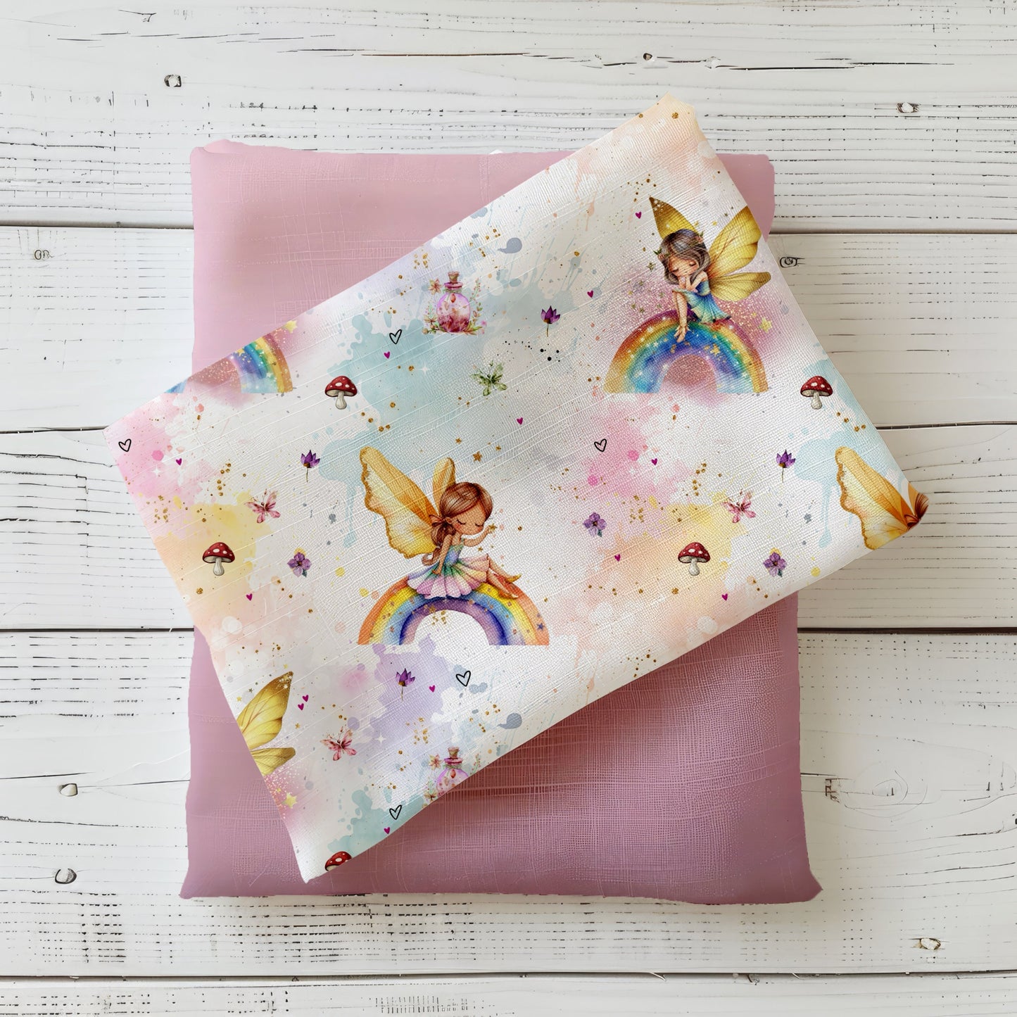 Gefalteter Kinderstoff mit Feen und Regenbogen-Motiven in Pastellfarben, dekorativ auf rosafarbenem Kombistoff präsentiert – ideal für Baby- und Kinderkleidung.
