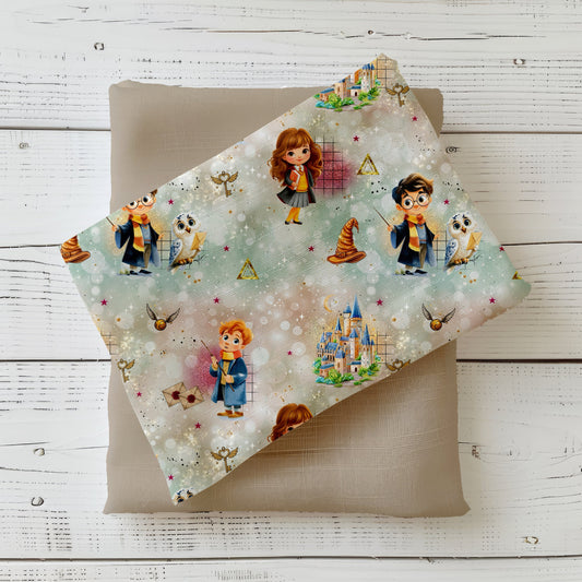 Mockup: Stoff „Zauberfreunde“ mit kleinen Zauberern/Zauberin, weißer Eule, Schloss, Briefen, goldener Kugel und Zauberhut auf pastelligem Bokeh-Hintergrund; aufgelegt auf taupefarbenem Kombistoff, fotografiert auf hellem Holz.