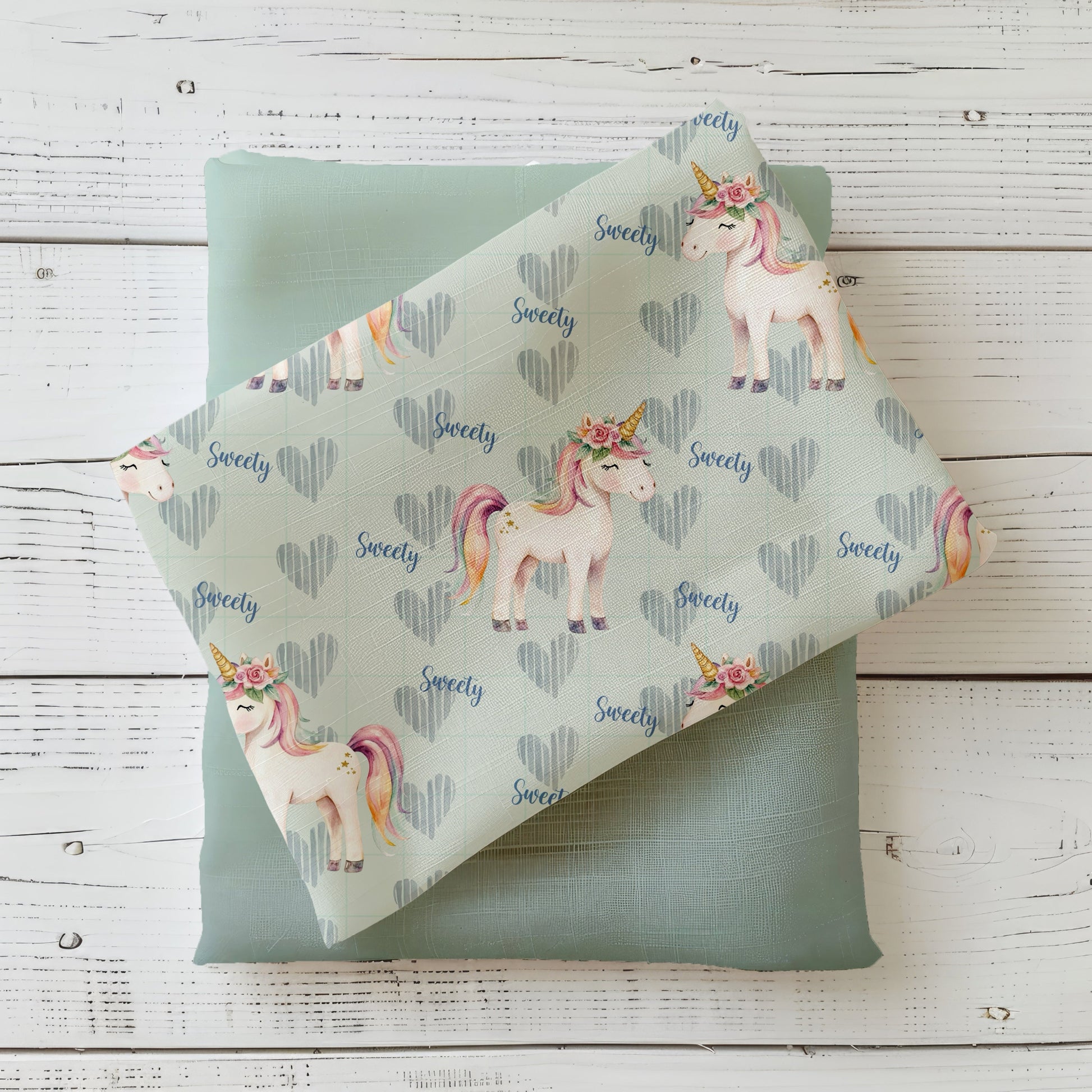 Mockup: Stoff „Zuckersüßes Einhorn“ – Einhörner mit Blumenkranz & Regenbogenmähne, Herzen im Karo und „Sweety“-Schrift auf mintfarbenem Hintergrund; Stoffkachel diagonal auf Uni in Salbei/Mint als Kombi.