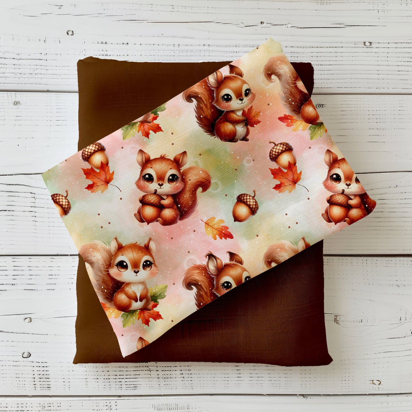 Gefalteter Kinderstoff mit herbstlichen Eichhörnchen, Eicheln und bunten Blättern auf pastelligem Hintergrund, dekorativ auf braunem Kombistoff präsentiert.
