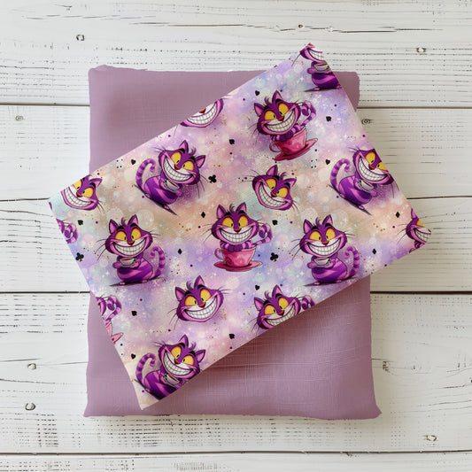 Mockup: Stoff „Der Grinsekater“ mit lila-pinker, breit grinsender Katze, Teetassen und Karten-Symbolen auf pastelligem Bokeh-Verlauf; der Druck liegt diagonal auf passendem Uni-Kombistoff.