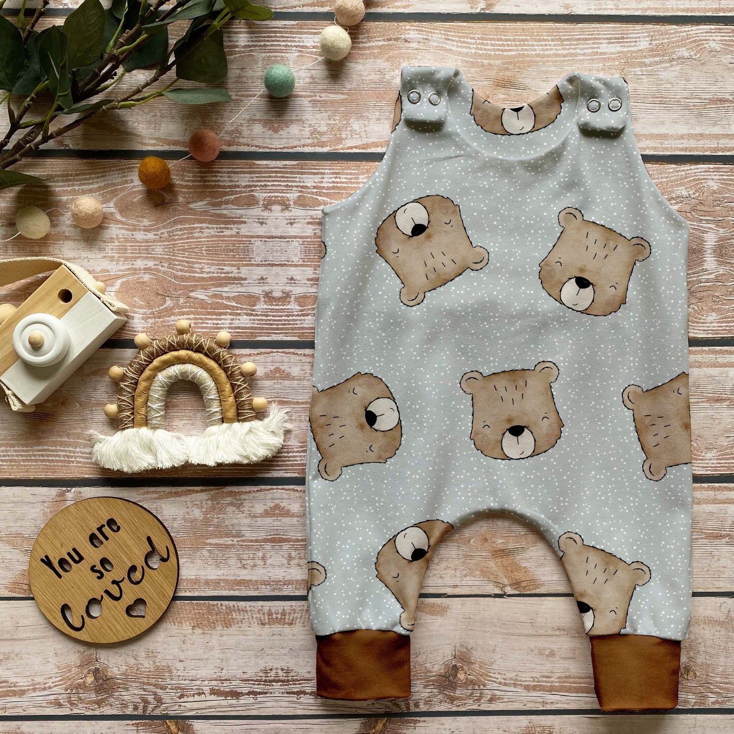 Handgemachter Baby-Strampler im Design „Benji der Bär“ mit süßen Bärenköpfen auf hellem Hintergrund und Bündchen in Karamell, liebevoll auf Holz mit naturbezogenen Dekoelementen inszeniert.