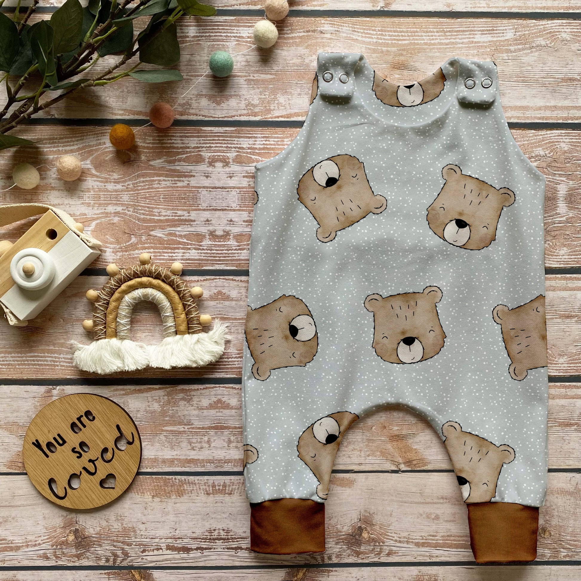Handgemachter Baby-Strampler im Design „Benji der Bär“ mit süßen Bärenköpfen auf hellem Hintergrund und Bündchen in Karamell, liebevoll auf Holz mit naturbezogenen Dekoelementen inszeniert.