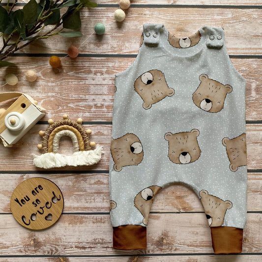 Handgemachter Baby-Strampler im Design „Benji der Bär“ mit süßen Bärenköpfen auf hellem Hintergrund und Bündchen in Karamell, liebevoll auf Holz mit naturbezogenen Dekoelementen inszeniert.