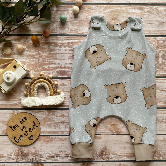 Handgemachter Baby-Strampler im Design „Benji der Bär“ mit süßen Bärenköpfen auf hellem Hintergrund und beigen Bündchen, liebevoll arrangiert auf Holzuntergrund mit Naturdeko.
