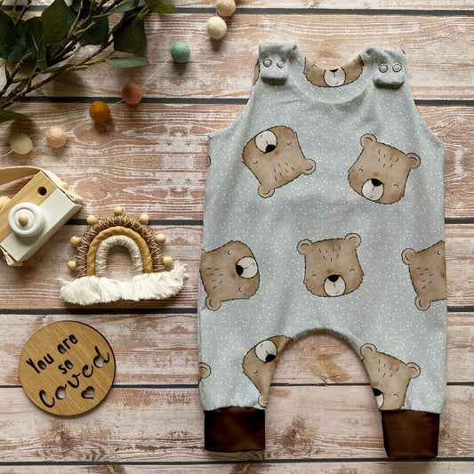 Handgemachter Baby-Strampler im Design „Benji der Bär“ mit süßen Bärenköpfen auf hellem Hintergrund und schokobraunen Bündchen, liebevoll dekoriert auf rustikalem Holzuntergrund.