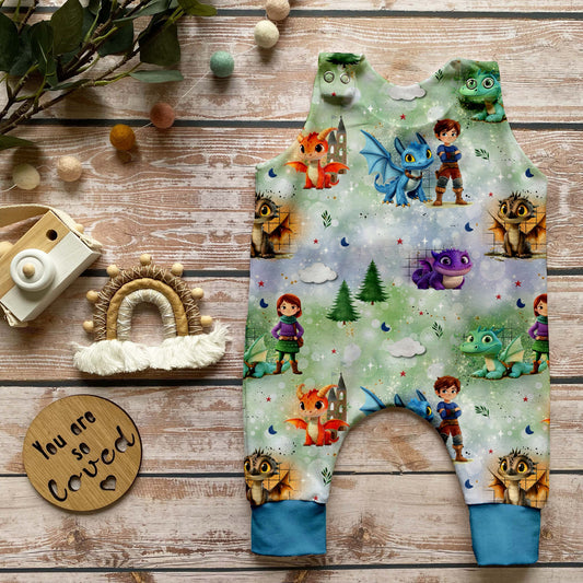 Handgemachter Baby-Strampler im Design „Die Drachenwächter“ mit bunten Drachenfiguren und Kindern auf zauberhaftem Hintergrund. Bündchen in frischem Azurblau, dekorativ arrangiert auf hellem Holzuntergrund mit Naturaccessoires.