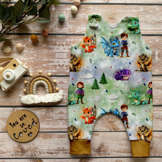 Handgemachter Baby-Strampler im Design „Die Drachenwächter“ mit kindlichen Figuren, Drachen und Burgen auf traumhaftem Hintergrund sowie Bündchen in dunklem Curry, liebevoll arrangiert auf hellem Holz mit Naturdeko.