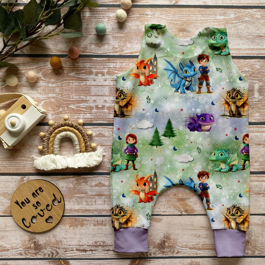 Handgemachter Baby-Strampler im Design „Die Drachenwächter“ mit kindgerechten Drachenmotiven und Figuren auf sanftem Hintergrund sowie fliederfarbenen Bündchen. Fotografiert auf hellem Holz mit liebevoller Naturdekoration.
