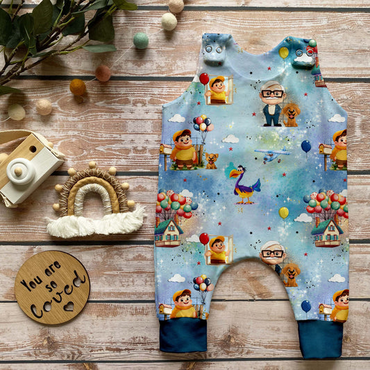 Handgemachter Baby-Strampler im Design „Fliegende Träume“ mit bunten Figuren, Luftballons, fliegenden Häusern und Tieren auf himmelblauem Hintergrund, ergänzt durch pfauenblaue Bündchen. Liebevoll inszeniert auf hellem Holz mit natürlichen Accessoires.