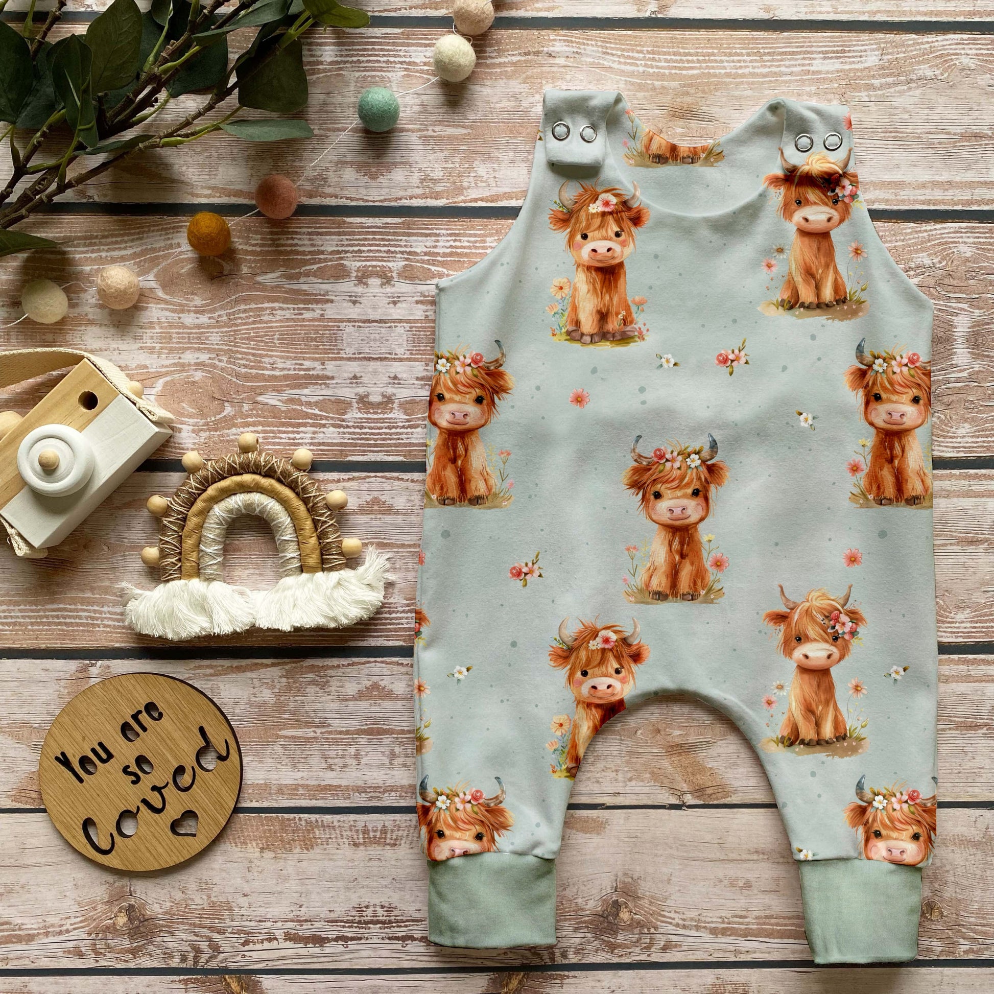 Handgemachter Baby-Strampler im Highland-Cow-Design mit niedlichen Kühen auf hellem Hintergrund und Bündchen in Schattengrün, liebevoll auf Holz mit Naturdeko inszeniert.