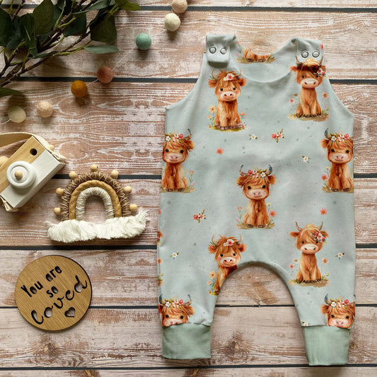 Handgemachter Baby-Strampler im Highland-Cow-Design mit niedlichen Kühen auf hellem Hintergrund und Bündchen in Schattengrün, liebevoll auf Holz mit Naturdeko inszeniert.