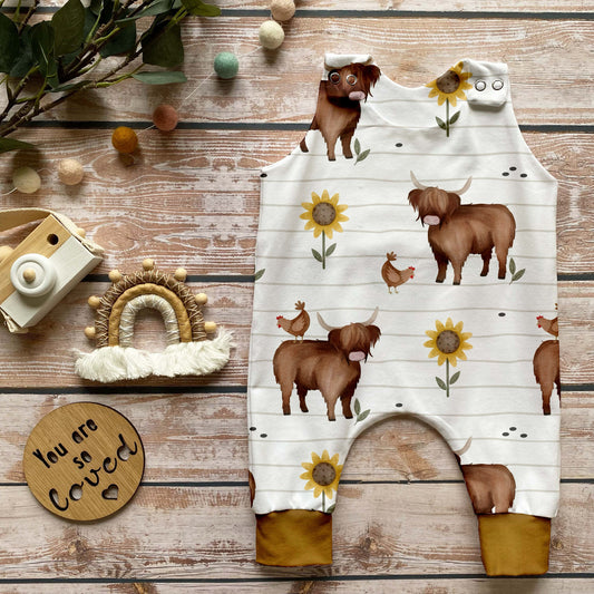 Handgemachter Baby-Strampler im Design „Hochlandrind“ mit Kühen, Hühnern und Sonnenblumen auf hellem Hintergrund und Bündchen in dunklem Curry, liebevoll fotografiert mit Holz-Deko.