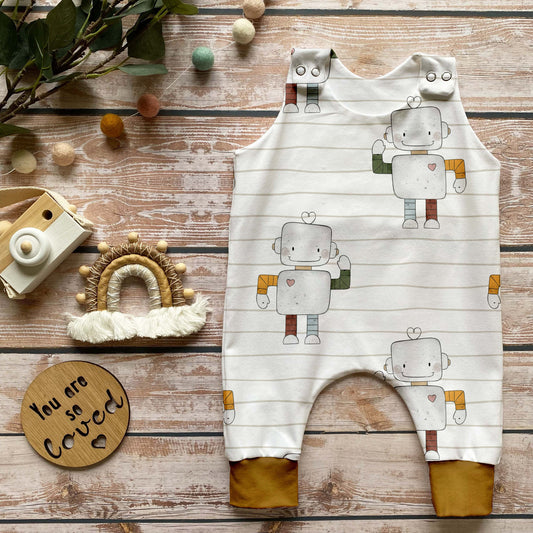 Handgemachter Baby-Strampler im Design „Kleiner Roboter“ mit freundlichen Robotern auf hell gestreiftem Hintergrund und Bündchen in dunklem Curry, liebevoll in Szene gesetzt mit Holzspielzeug.
