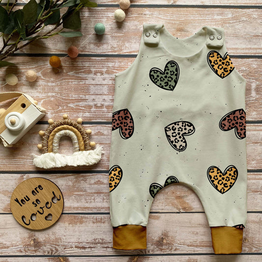 Handgemachter Baby-Strampler im Design „Leo Herzen“ mit herzförmigem Leo-Muster auf hellem Hintergrund und curryfarbenen Bündchen, liebevoll inszeniert auf Holz mit Dekoelementen.