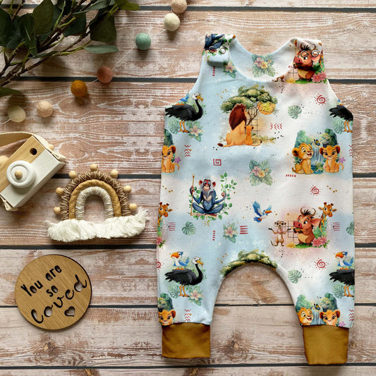 Handgemachter Baby-Strampler im Design „Savannenkönig“ mit fröhlichen Tierszenen, afrikanischen Naturmotiven und Bündchen in dunklem Curry, arrangiert auf hellem Holzuntergrund mit Deko aus Naturmaterialien.