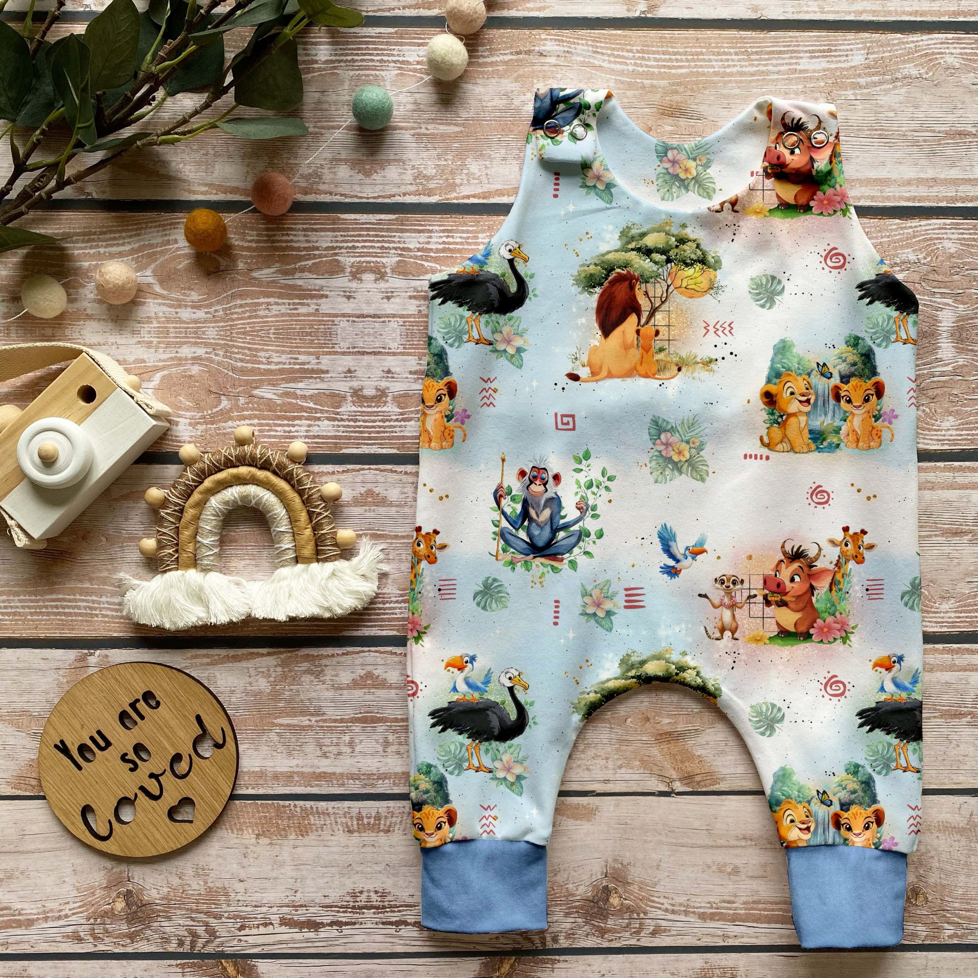 Handgemachter Baby-Strampler im Design „Savannenkönig“ mit liebevoll gezeichneten Tieren und Naturmotiven auf hellem Hintergrund sowie zart eisblauen Bündchen. Arrangiert auf Holzuntergrund mit natürlicher Deko.