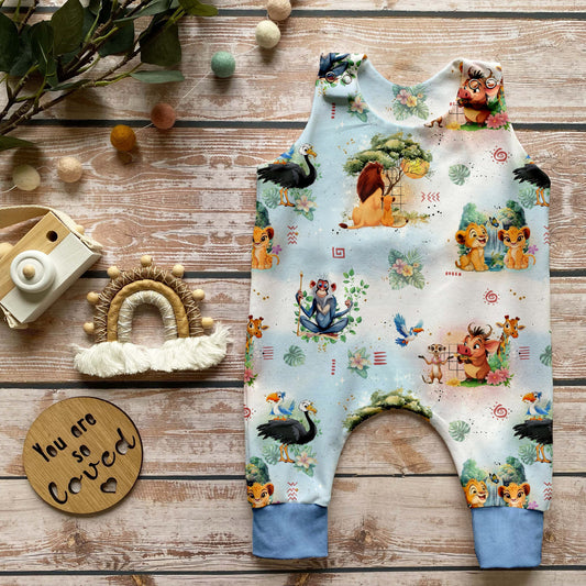 Handgemachter Baby-Strampler im Design „Savannenkönig“ mit liebevoll gezeichneten Tieren und Naturmotiven auf hellem Hintergrund sowie zart eisblauen Bündchen. Arrangiert auf Holzuntergrund mit natürlicher Deko.