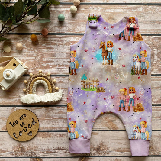 Handgemachter Baby-Strampler im Design „Zauberhafte Reiterinnen“ mit Reiterinnen, Pferden und Naturmotiven auf zartem Hintergrund sowie lavendelfarbenen Bündchen, arrangiert auf hellem Holz mit natürlichen Accessoires.