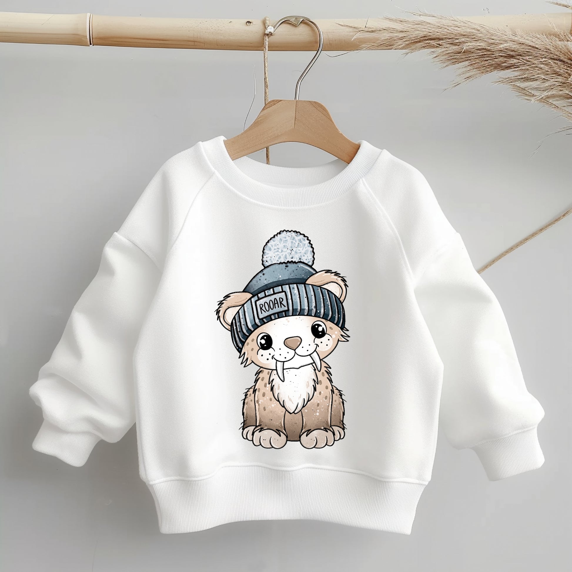 Freigestelltes Bügelbild „Säbi der Säbelzahntiger mit Mütze“ – klare Kontur, warme Naturtöne, blaue Mütze; ideal als Aufbügelmotiv für Kinder-Shirts, Hoodies, Mützen oder Taschen.